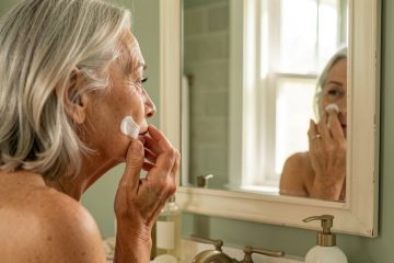 Femme de 67 ans aux cheveux argentés appliquant délicatement une crème hydratante sur son visage dans une salle de bains lumineuse.